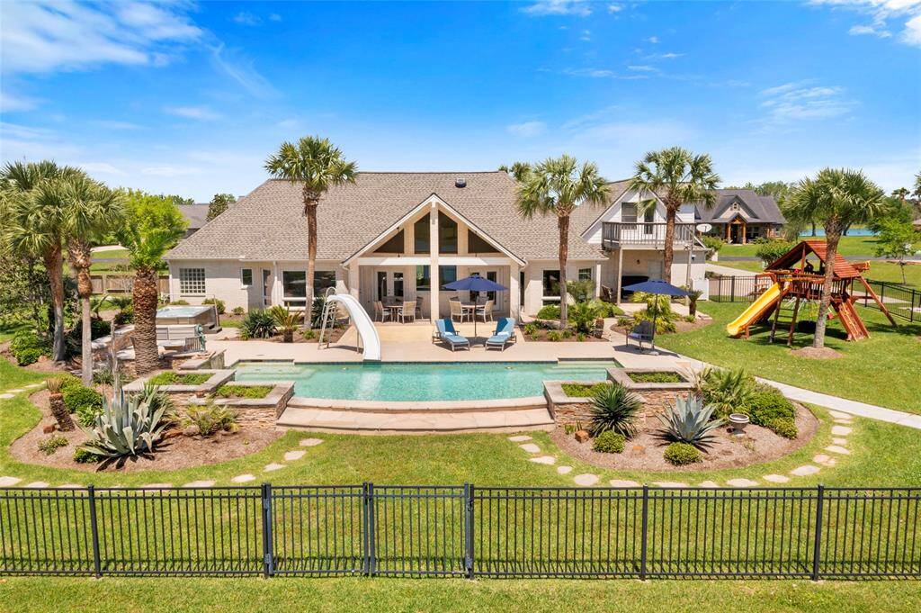 This spacious backyard has a large pool and waterslide, surrounded by lush landscaping and palm trees. There’s a play area for children and ample outdoor seating, perfect for entertaining. The home’s exterior boasts a modern design with large windows and a welcoming covered back patio.