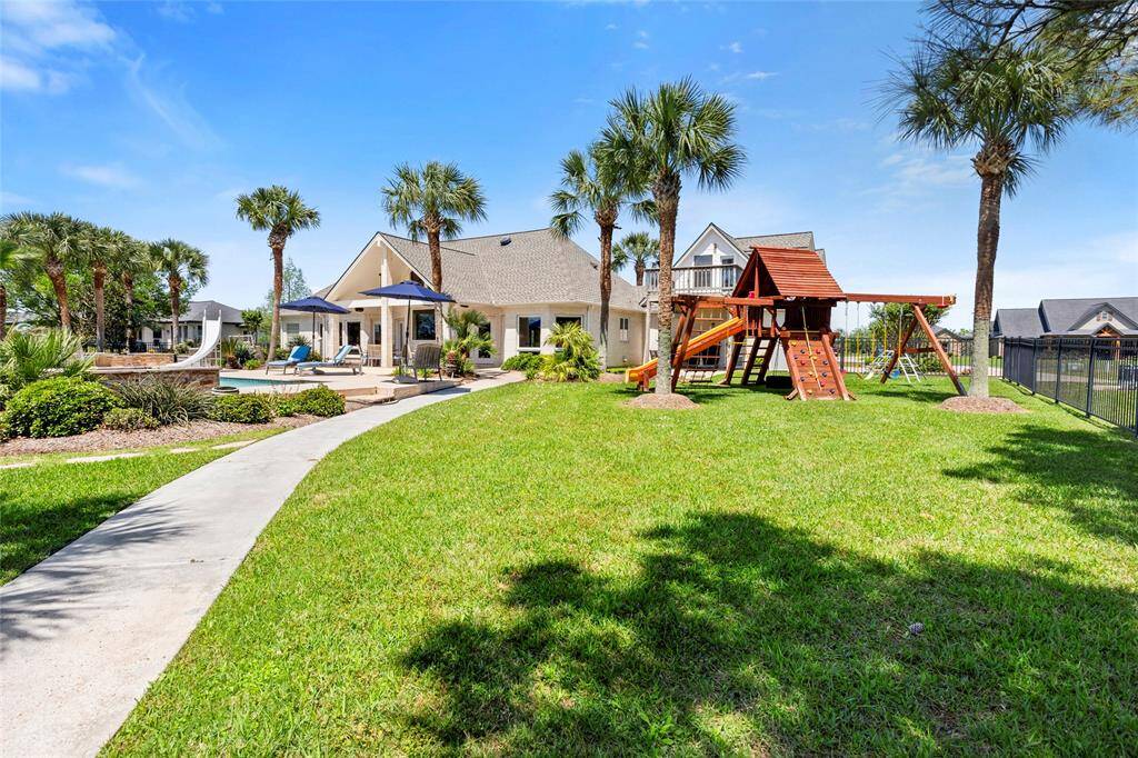 Escape to your tropical oasis with a lush lawn, children's playset, and a paved path leading to a poolside patio with seating and umbrellas, surrounded by beautiful palm trees.