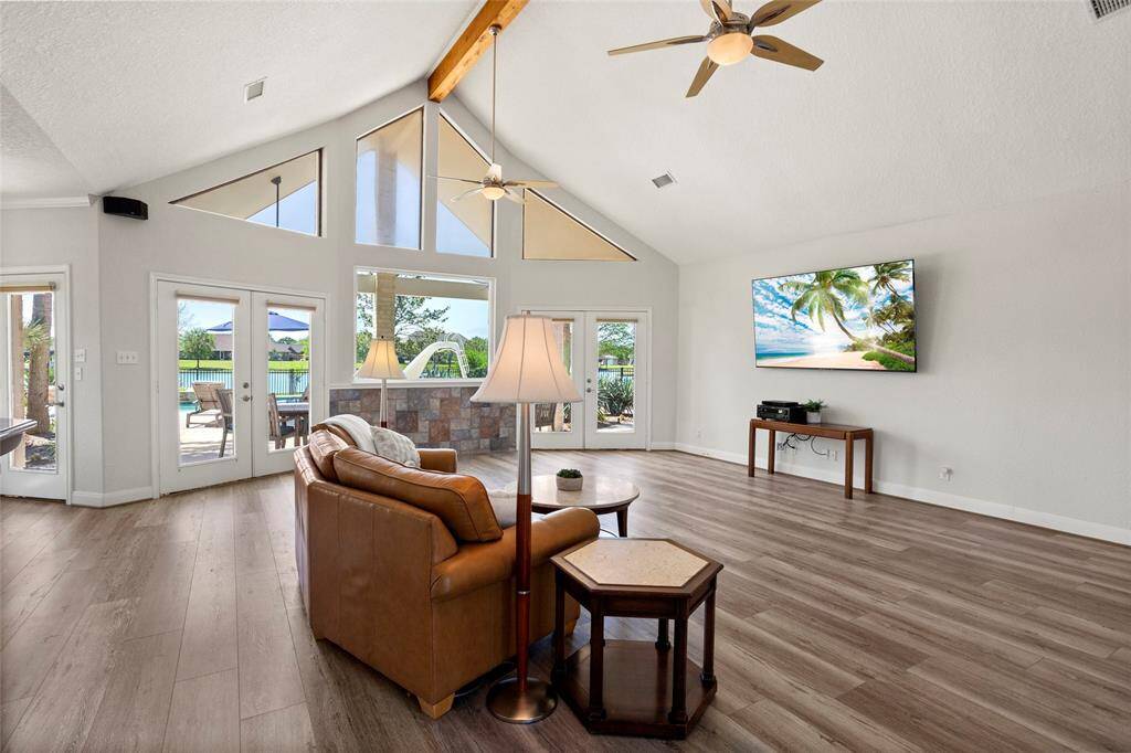 This inviting living room boasts vaulted ceilings with a wooden beam, modern fans, and floor-to-ceiling windows for abundant natural light. French doors open to a covered patio with pool views, complemented by sleek wood LVP flooring.