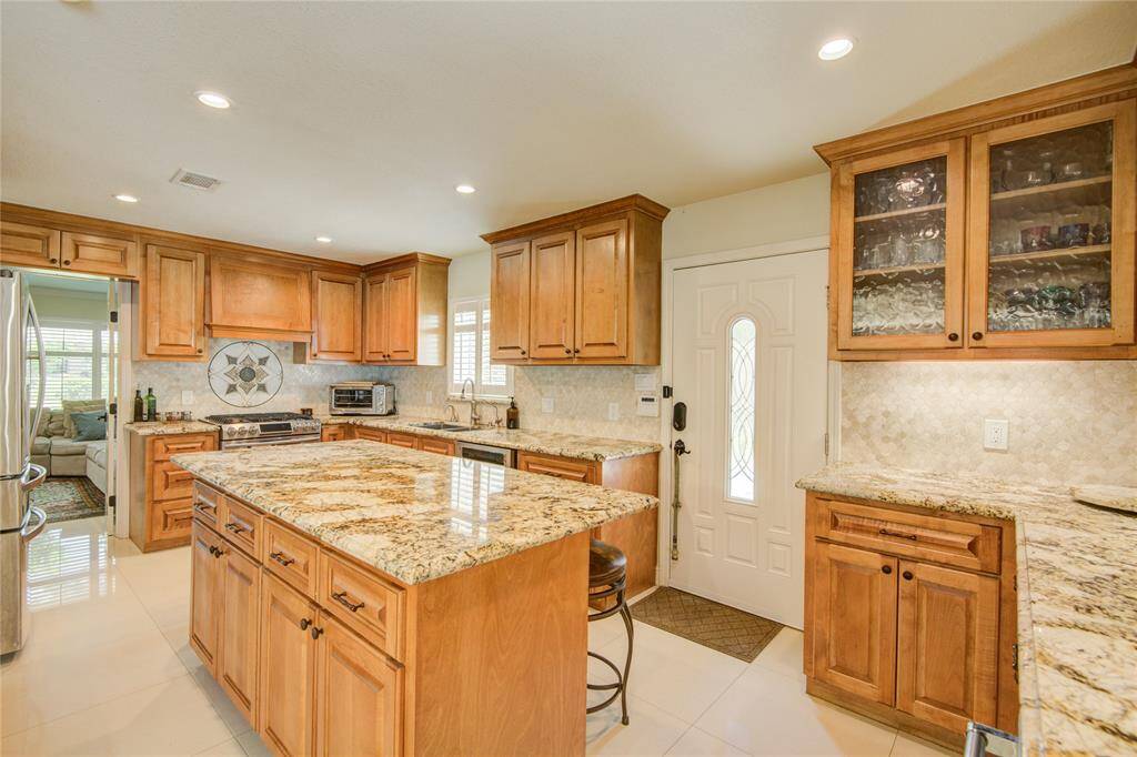 The kitchen boasts plentiful soft-close wood cabinetry, stainless steel appliances, a spacious granite island with a belly bar, and a glimpse of the formal living room to the left.