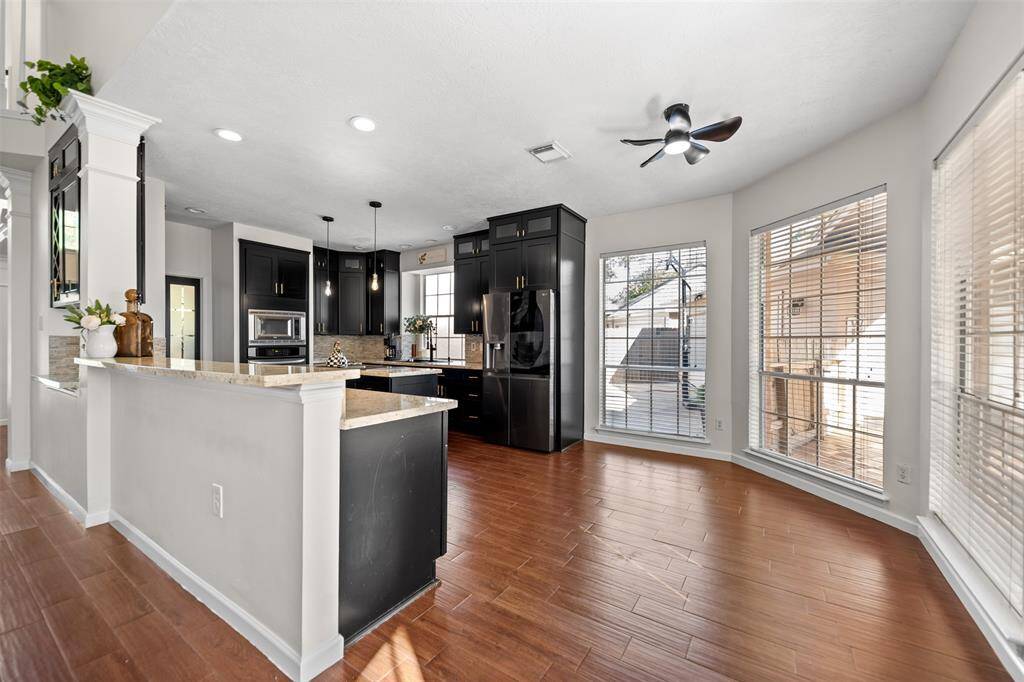 Bright kitchen with modern black cabinetry, stainless steel appliances, and ample counter space. The area features large windows, wood-style flooring, and a ceiling fan, creating a welcoming and functional space.