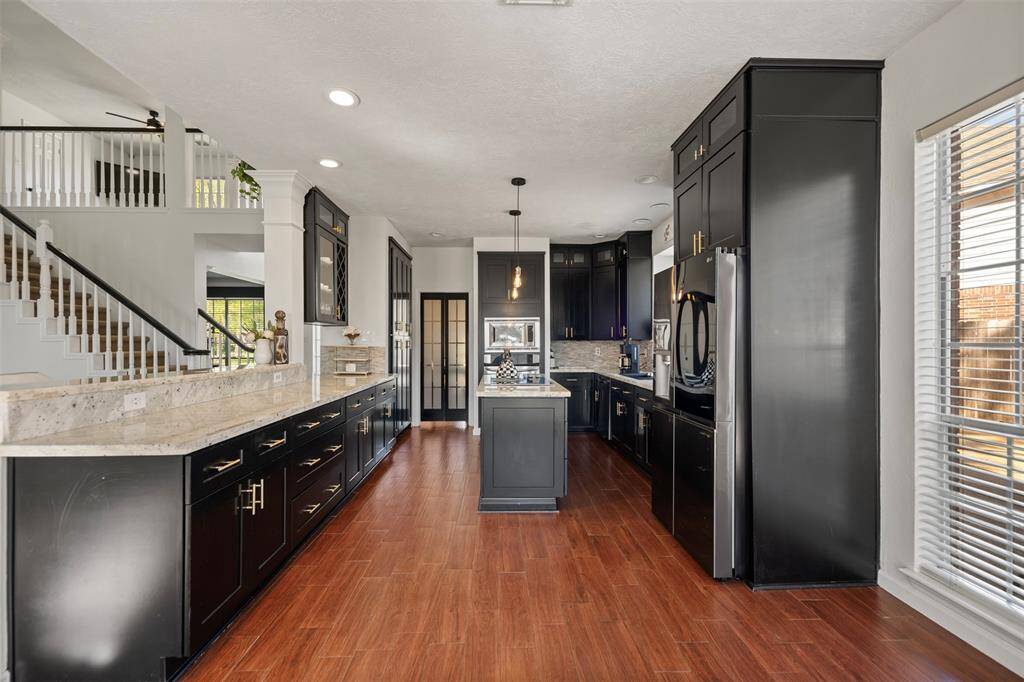 This modern kitchen features sleek black cabinetry, stainless steel appliances, and a spacious marble countertop. The open layout includes a view of the staircase and large windows for natural light, complemented by warm wood flooring.