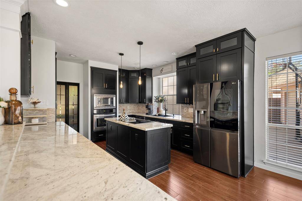 This modern kitchen features sleek black cabinetry, stainless steel appliances, and a central island with pendant lighting. The space is enhanced by large windows, offering ample natural light, and it has elegant wood flooring.