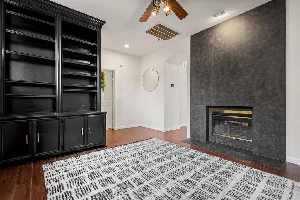 This cozy living space features a modern double-sided fireplace with a sleek, dark surround and a built-in black bookshelf. The room has warm wood flooring, a patterned area rug, and a ceiling fan for comfort. Ideal for relaxation and stylish storage or office space.