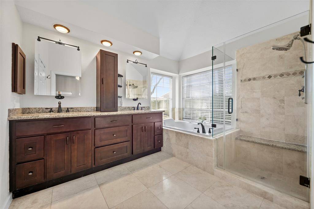 This spacious bathroom features a double vanity with granite countertops, a large soaking tub, and a glass-enclosed shower. Natural light floods in through the corner windows, highlighting the elegant tile flooring and modern fixtures.