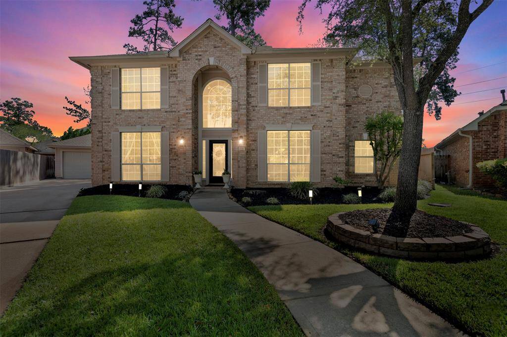 This photo showcases a charming two-story brick house with large windows, beautifully lit at dusk. The well-maintained front yard features a manicured lawn, a curved walkway, and mature trees, creating an inviting curb appeal.