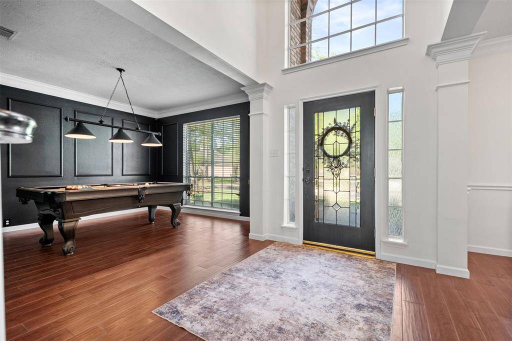 This photo showcases a stylish entrance with a modern glass door and sidelights, leading into a bright room featuring a pool table. The area has elegant dark accent walls, large windows for natural light, and warm wood flooring, offering a welcoming and entertainment-friendly space.