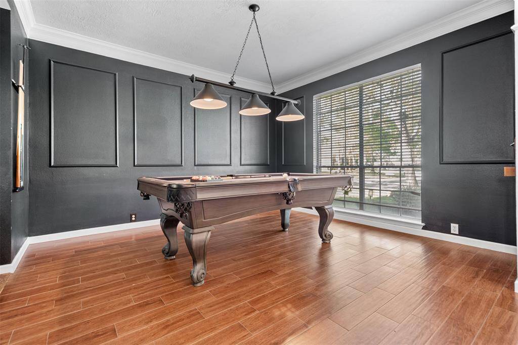 This photo shows a stylish room with a pool table, featuring dark paneled walls and wood-style flooring. The large window provides ample natural light, and overhead lighting adds a modern touch with a hinden bar under the staircase.