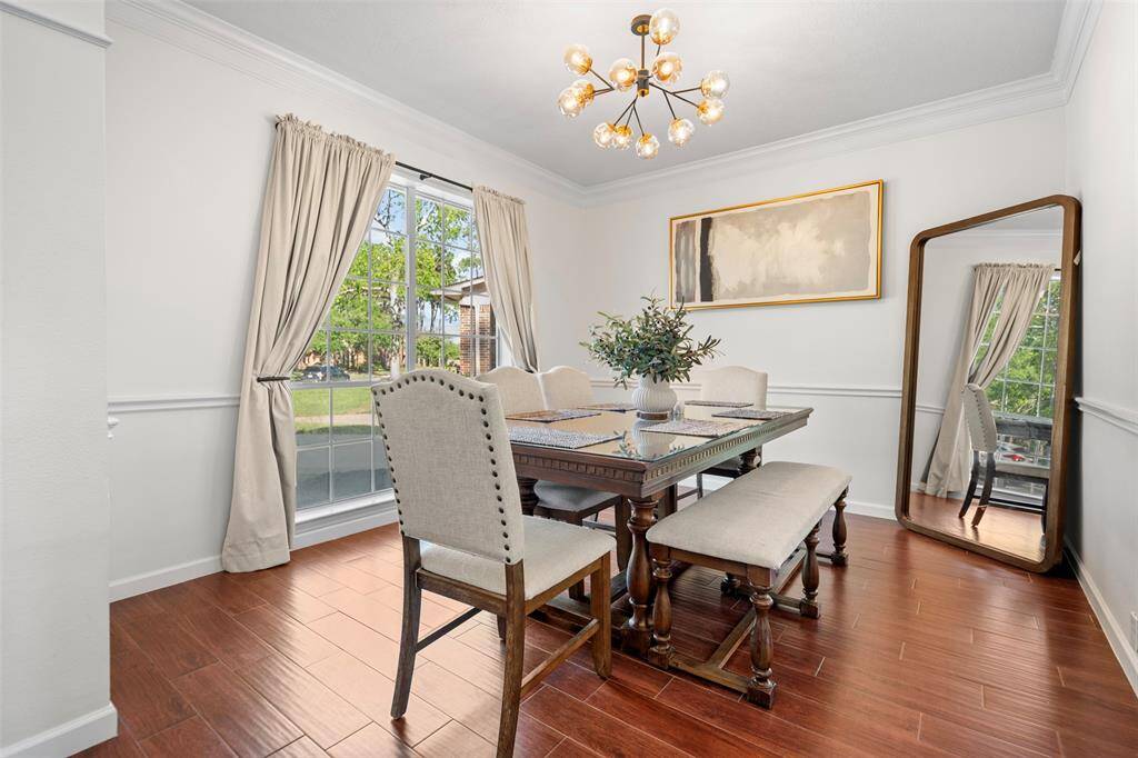 This dining room features elegant hardwood flooring, a stylish chandelier, and large windows with drapes that provide ample natural light. The space includes a classic dining table with upholstered chairs and a bench, complemented by a large mirror and tasteful artwork.
