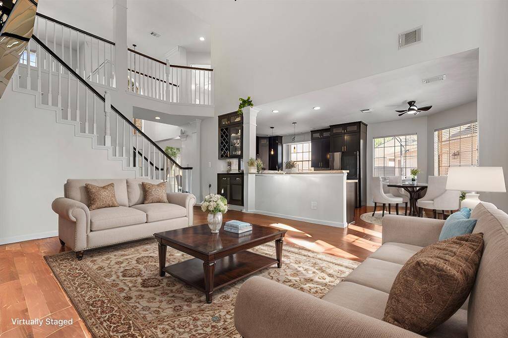 This photo showcases a bright, open-concept living area with a high ceiling and elegant staircase. The space features a cozy seating arrangement, hardwood floors, and a modern kitchen with dark cabinets and stainless steel appliances. Large windows provide ample natural light, enhancing the welcoming atmosphere. (Virtually Staged)