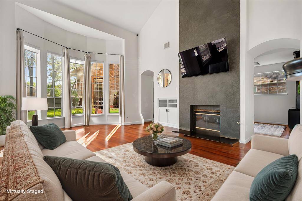 The living room is spacious with high ceilings and large windows offering abundant natural light. It features a modern fireplace, hardwood floors, and is tastefully staged with comfortable seating, a round coffee table, and a large TV. (Virtually Staged)