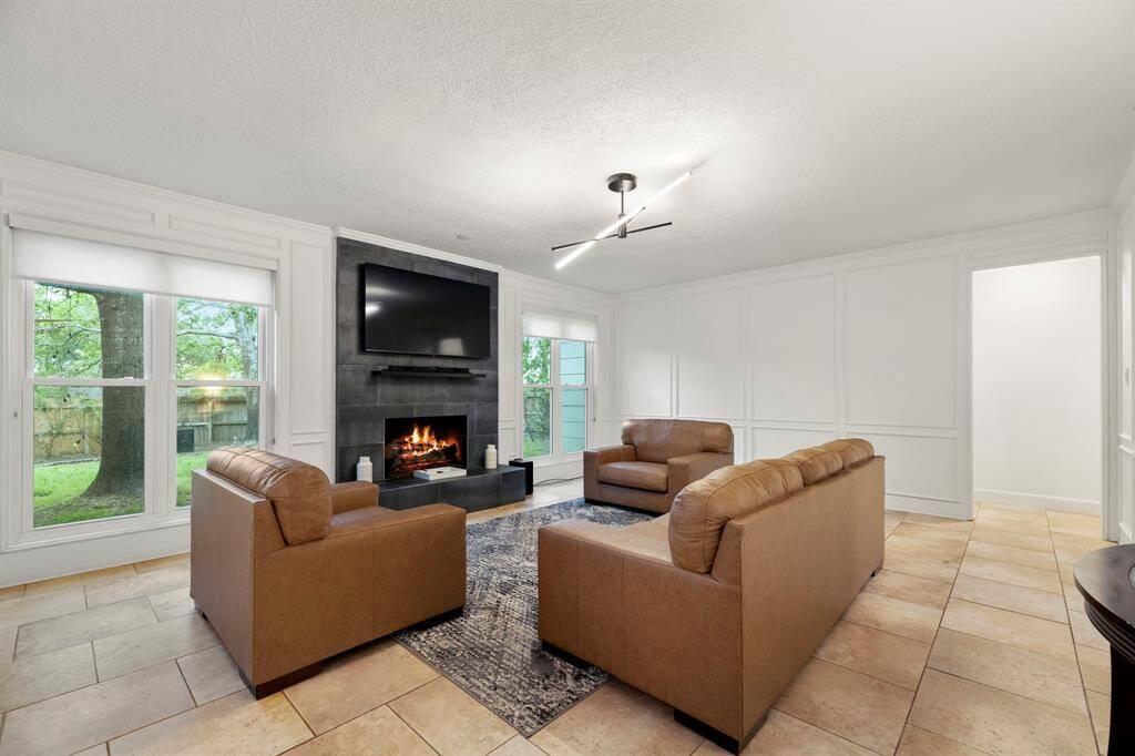 The living room features a cozy gas log fireplace surrounded by painted wood panel walls with picture frame molding, adding character and warmth to the space. Large windows overlook the backyard, bringing in plenty of natural light. The doorway to the right leads to the primary suite, providing easy access to your private retreat.