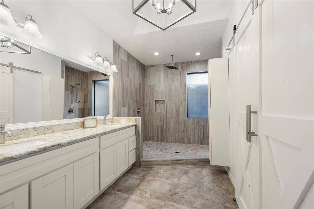 The primary bath features dual sinks with plenty of counter space to the left, offering ample room for your daily essentials. Ahead, a massive walk-in shower provides a luxurious experience. To the right, sliding barn doors open to reveal a spacious walk-in closet, providing plenty of storage for your wardrobe.