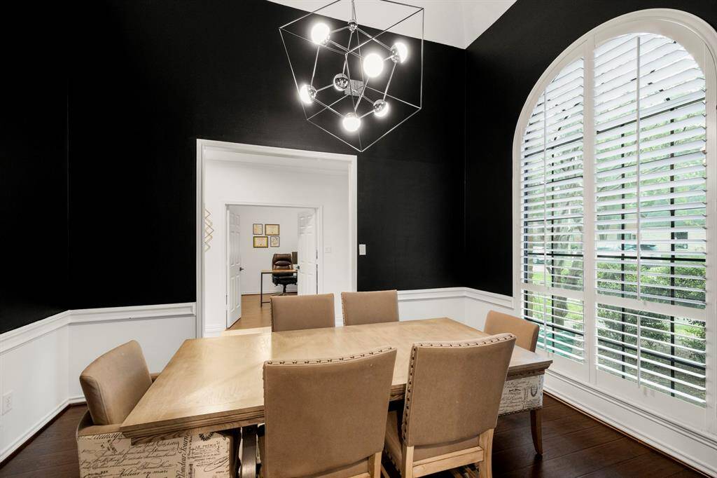 The formal dining room offers a sophisticated setting with plantation shutters on the windows overlooking the front yard. Raised ceilings, a modern chandelier, and chair railing enhance the space, while wood floors provide warmth throughout. Through the opening by the front door, you can glimpse the home office, creating a seamless flow between the rooms.