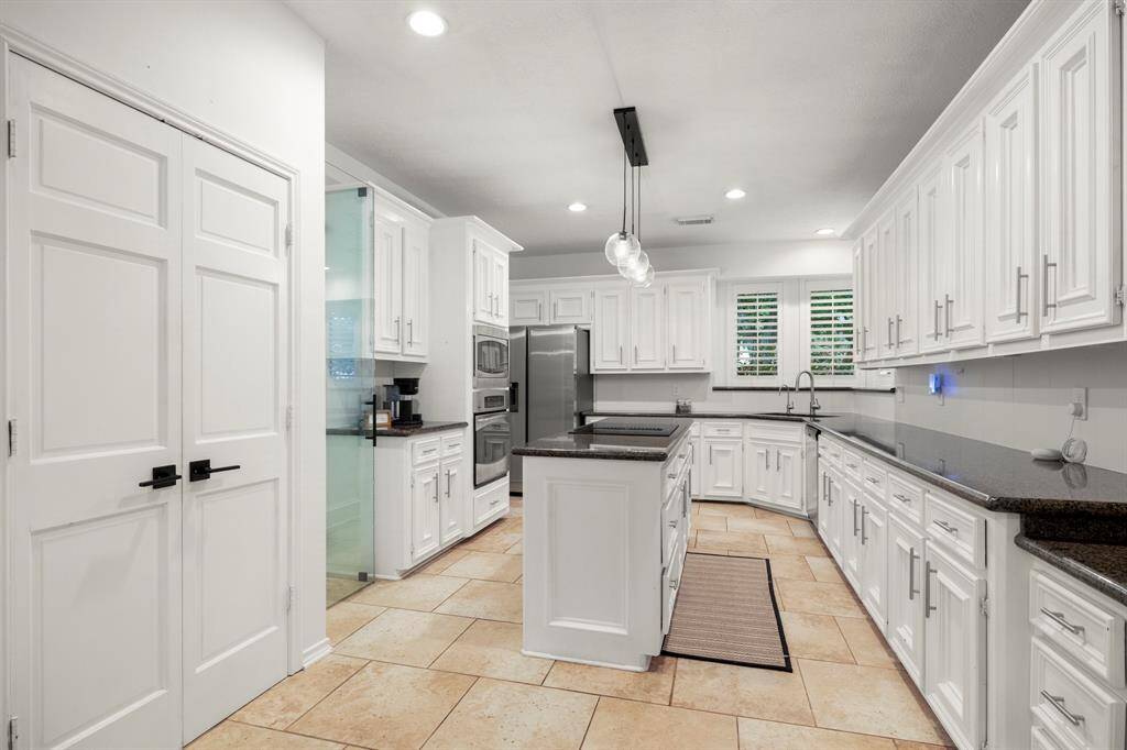 This kitchen is a dream with abundant cabinet space and plenty of storage! Bright cabinets complement gleaming granite countertops, while a modern light fixture hangs over the island. Elegant trim and plantation shutters on the windows above the corner sink add a refined touch. Stainless appliances, a glass cooktop in the island, and a stylish glass-enclosed wine storage complete the space. The doorway to the formal dining room is just out of sight by the fridge, offering convenient access.