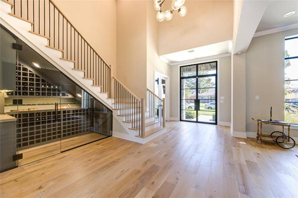 This photo showcases a spacious, modern entryway with high ceilings and abundant natural light. It features a sleek staircase with a glass-enclosed wine storage area underneath. The flooring is light wood, and the entrance has large windows and a stylish chandelier.