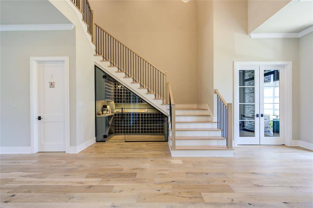 This photo features a spacious, modern entryway with light wood flooring and soft beige walls. A sleek staircase with metal railings leads to an upper level, and there's a wine storage area beneath it. French doors on the right open to a bright adjacent study room, adding to the open, inviting feel.