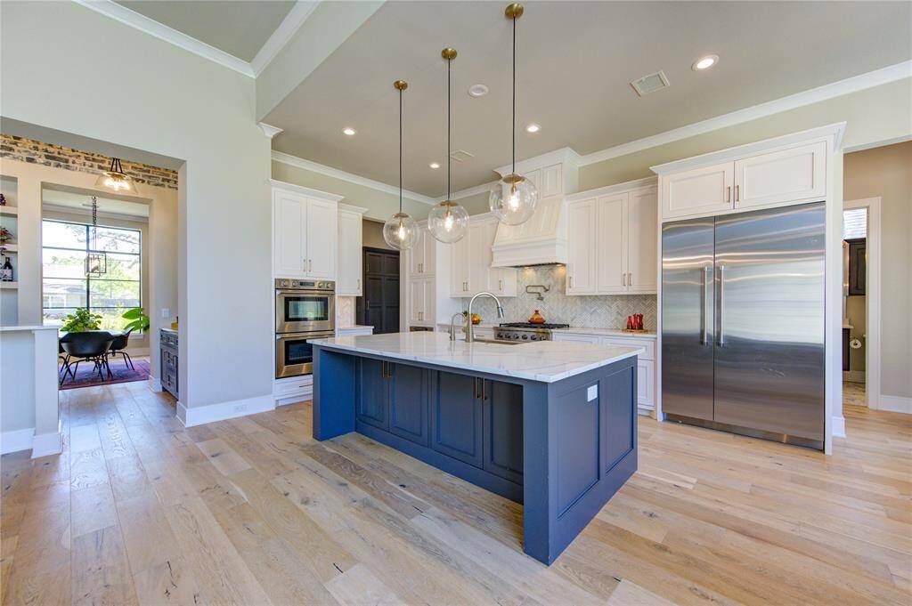 This photo showcases a modern kitchen with a large island featuring a white countertop and dark blue base. It has stainless steel appliances, white cabinetry, pendant lighting, and hardwood floors. An adjacent dining area with large windows is visible through an open doorway.