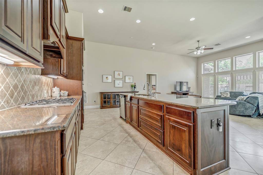 This ample open-concept kitchen and living area features a large island with a sink, elegant dark wood cabinetry, and tiled floors. The living room is brightened by large windows, allowing plenty of natural light, and includes recessed lighting and a ceiling fan.
