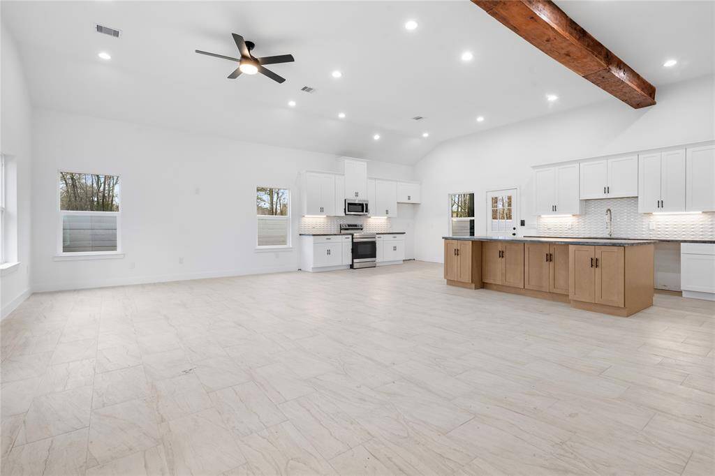 Modern open-concept kitchen and living area. It features light-toned tile flooring, white cabinets, stainless steel appliances, and a central island with wood accents. The space is well-lit with recessed lighting and a ceiling fan, complemented by large windows and a rustic wooden ceiling beam.