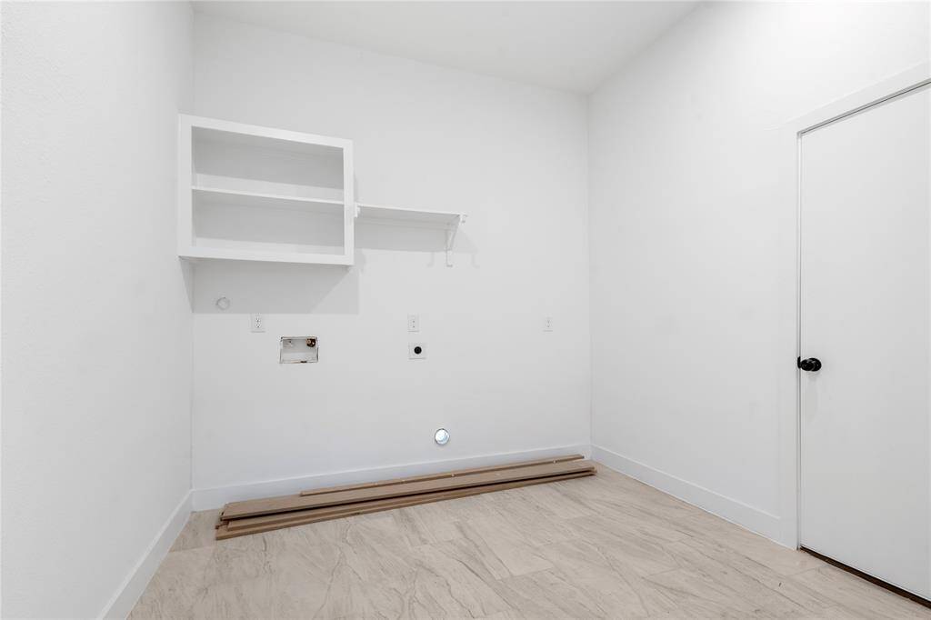 This is a bright, unfurnished utility room with a white wall, open shelving, and hookups for a washer and dryer. The space features light-colored flooring and a door for entry.