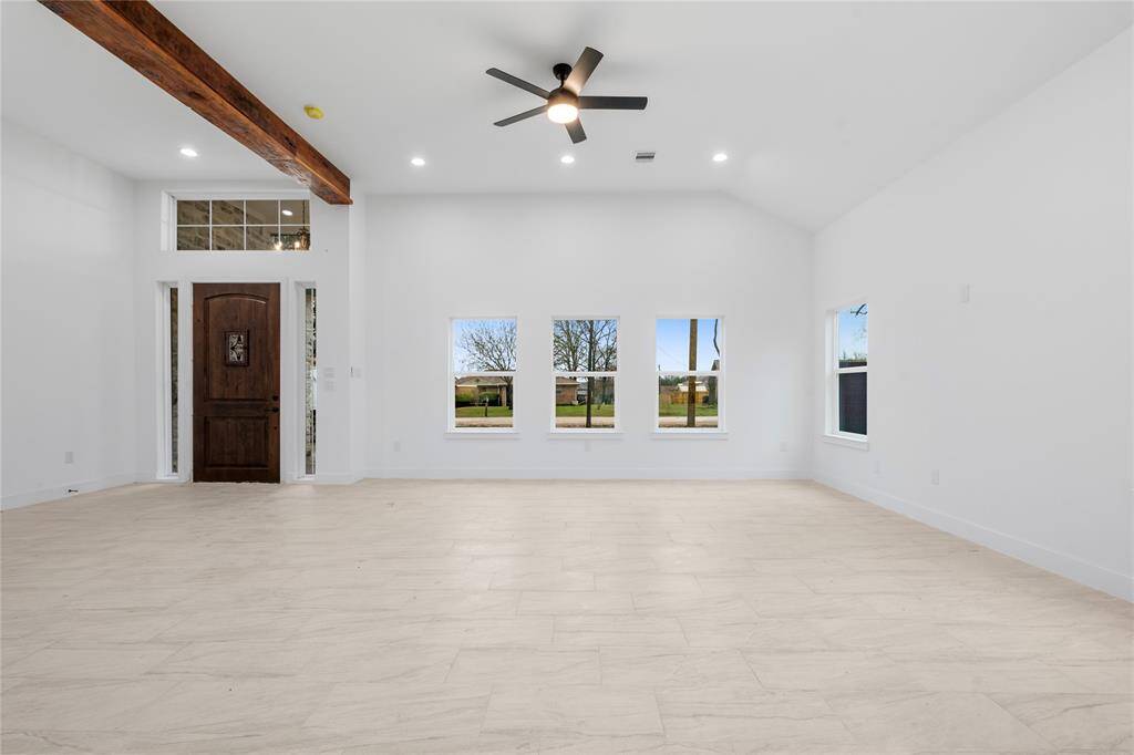 Bright, spacious living room with high ceilings, wood beam accents, and a modern ceiling fan. Features large windows for natural light and a stylish wooden front door.