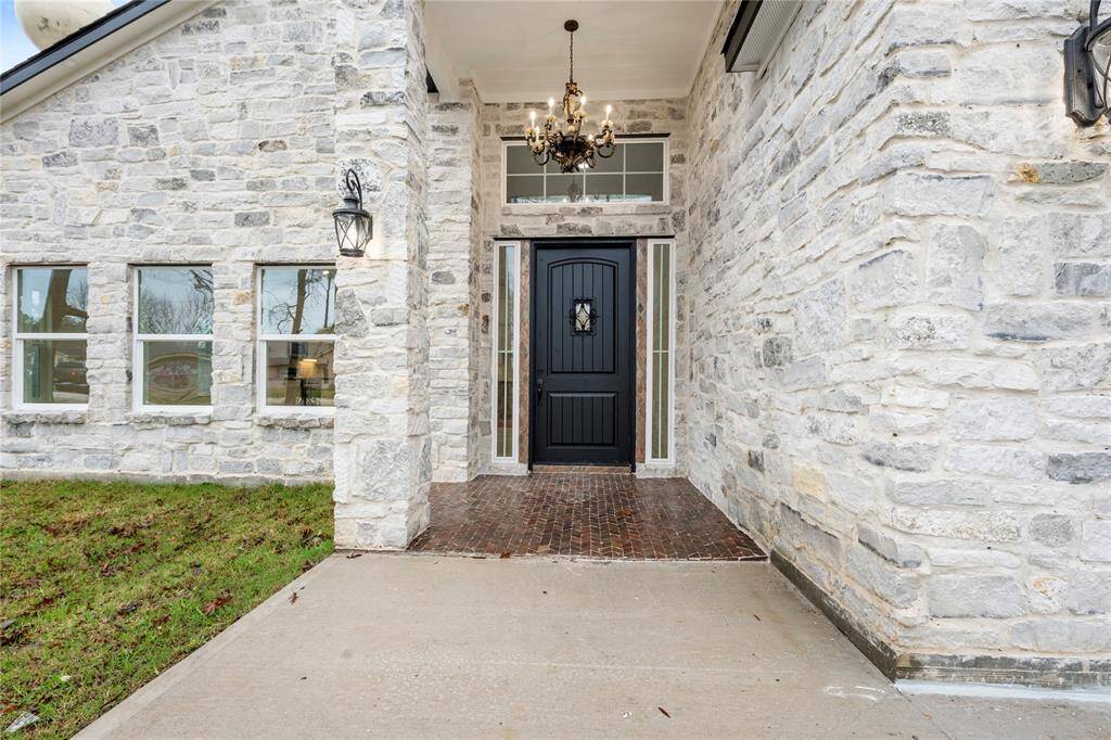 Charming stone-front entryway with a black door and side windows. It features a brick walkway, elegant outdoor lighting, and a decorative chandelier, offering a welcoming and stylish entrance to the home.