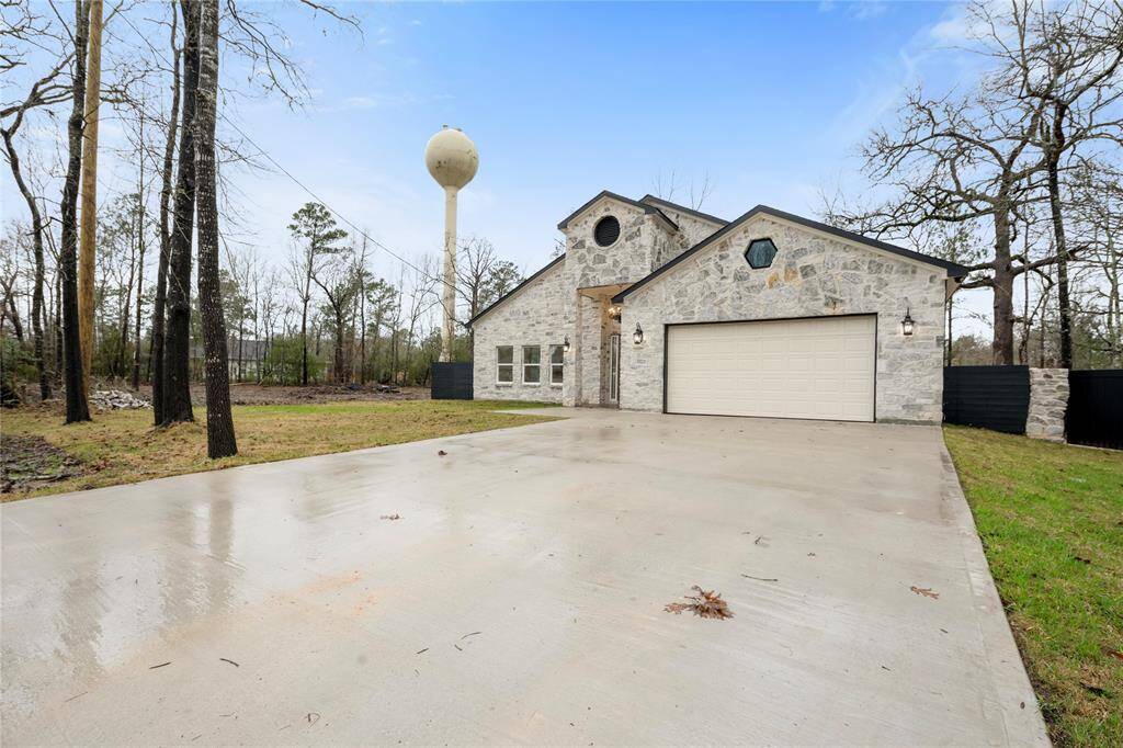Modern stone house with a two-car garage and a spacious driveway. It is surrounded by a wooded area, offering privacy and a natural setting. The exterior features a mix of rustic and contemporary design elements.