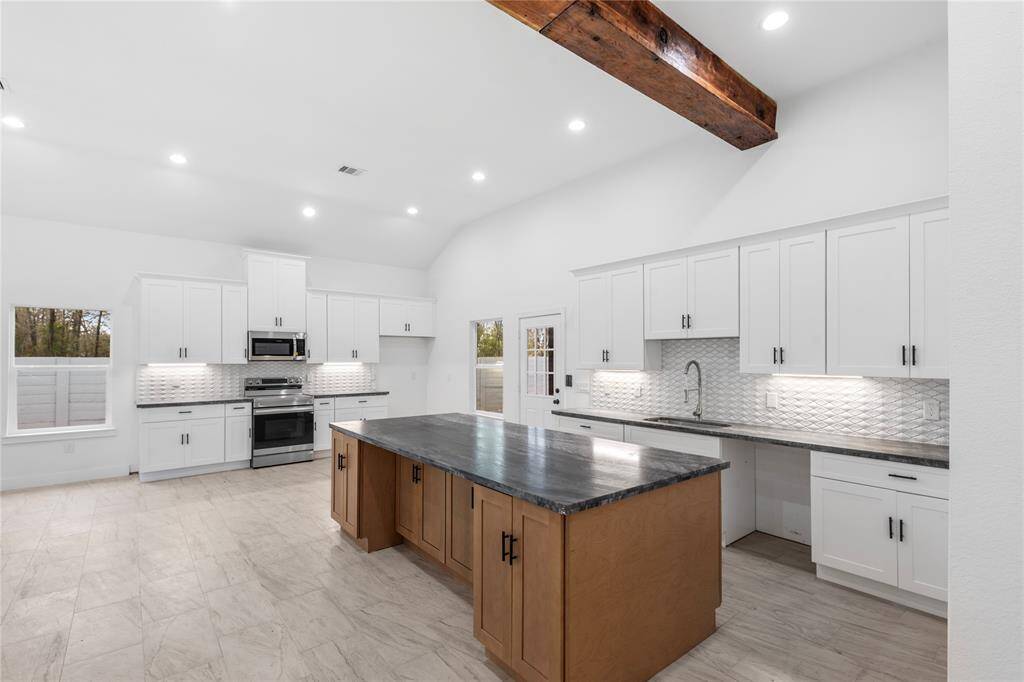 This kitchen features a spacious, open layout with modern white cabinetry, stainless steel appliances, and a large island with a dark countertop. The room has high ceilings with a rustic wooden beam and ample natural light from large windows, creating a bright and inviting space.