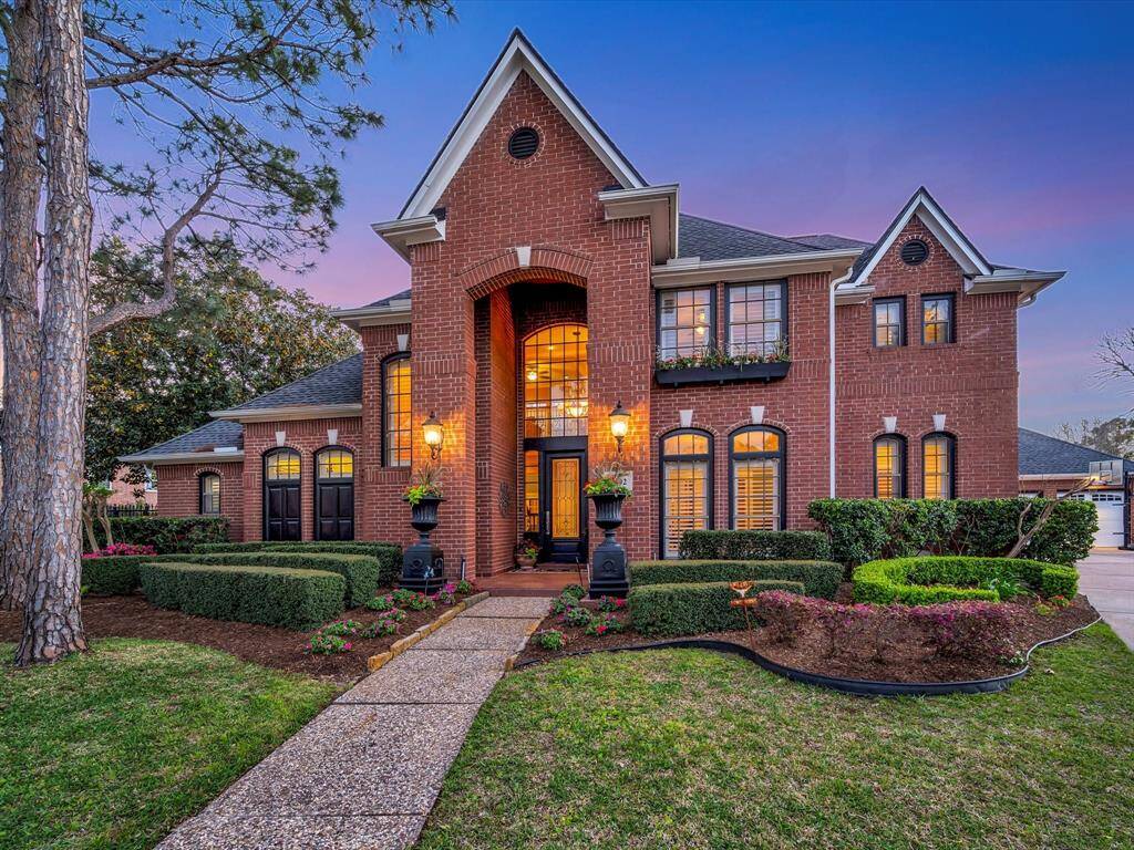 This stunning two-story brick home features a grand entrance with large windows and elegant landscaping. A well-maintained pathway leads to the front door, flanked by charming lanterns. The house is surrounded by lush greenery, offering a welcoming and luxurious curb appeal.
