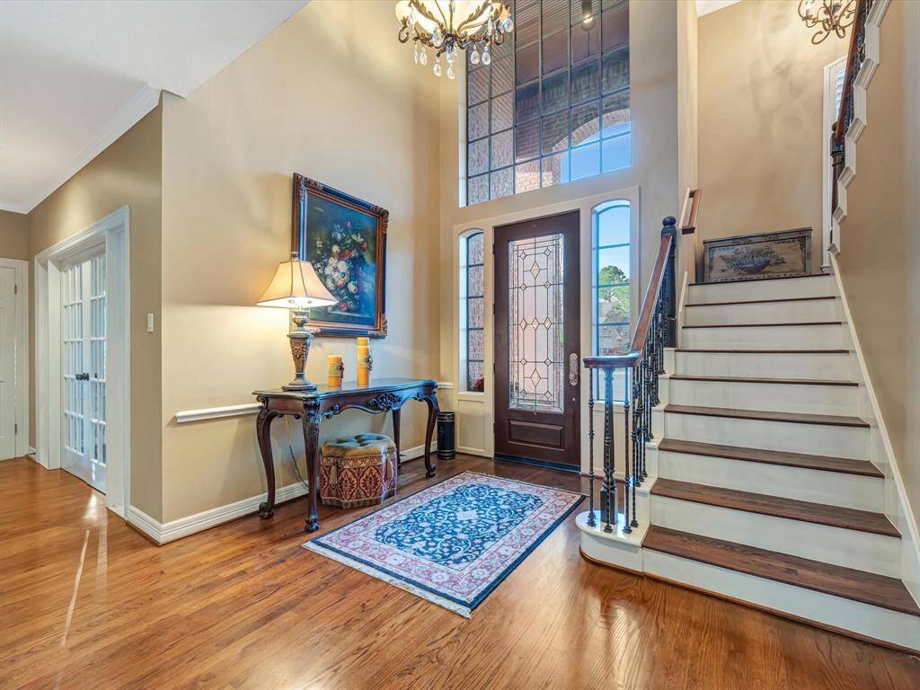 This photo showcases an elegant entryway with a high ceiling, large windows, and a stylish chandelier. The space features a beautiful staircase with wrought iron railings, hardwood floors, and a decorative rug. A console table with a lamp and artwork adds a welcoming touch.