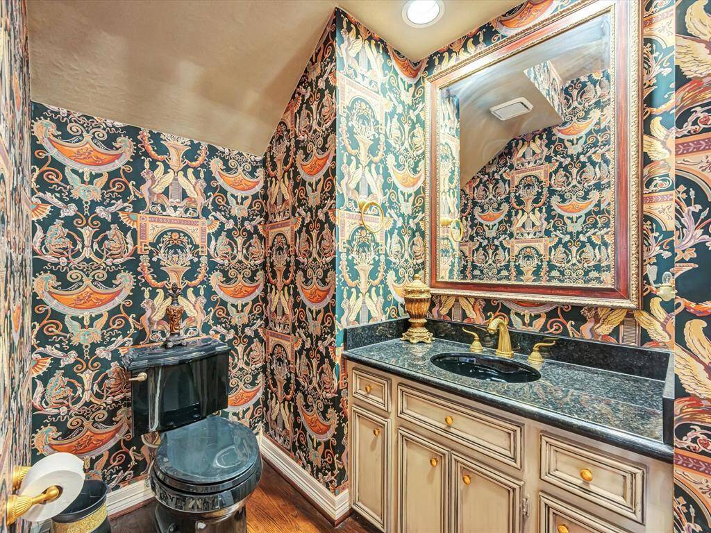 This bathroom features bold, ornate wallpaper with a traditional design, complementing a black granite countertop and matching sink. The cabinetry has a distressed finish, and gold fixtures add a touch of elegance. A large framed mirror enhances the space, making it feel more open and sophisticated.
