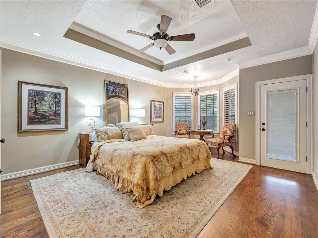 This spacious bedroom features elegant tray ceilings, a ceiling fan, and warm wood flooring. The room is well-lit with natural light from large windows with shutters, and it includes a cozy sitting area. A door provides direct outdoor access.