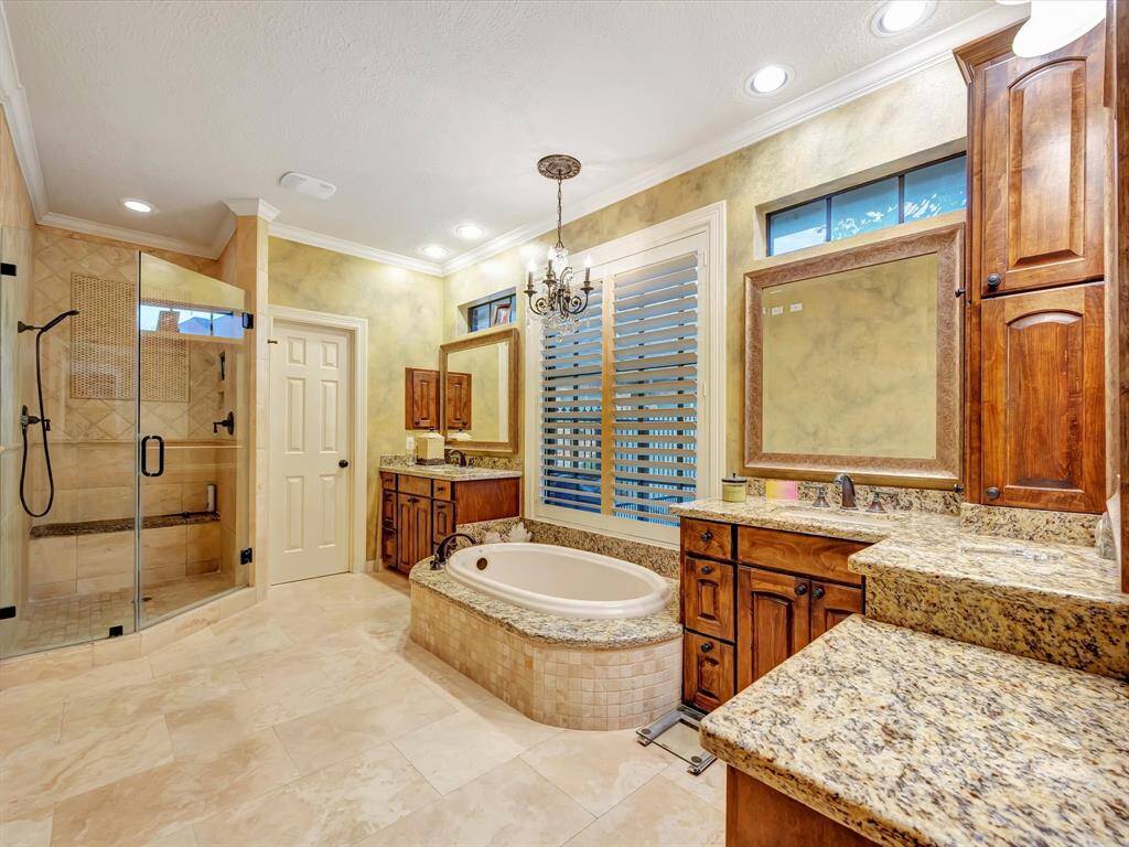 This luxurious bathroom features a spacious layout with a large glass-enclosed shower, a deep soaking tub, and elegant granite countertops. Warm wood cabinetry and a chandelier add a touch of sophistication, while ample natural light creates an inviting atmosphere.