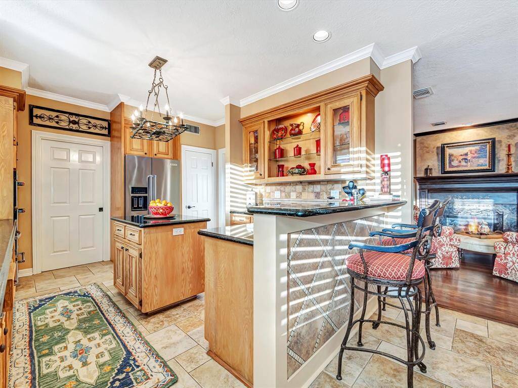 This inviting kitchen features warm wood cabinetry, granite countertops, and a central island with a hanging pot rack. The space includes a breakfast bar with stylish seating, tile flooring, and a cozy view into the living area with a fireplace. Ideal for entertaining and everyday living.