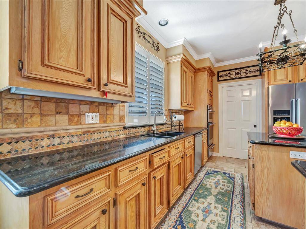 This kitchen features warm wood cabinetry, granite countertops, and a decorative tile backsplash. It includes stainless steel appliances and a central island with a hanging pot rack. The space is bright and inviting with a cozy runner on the tile floor.