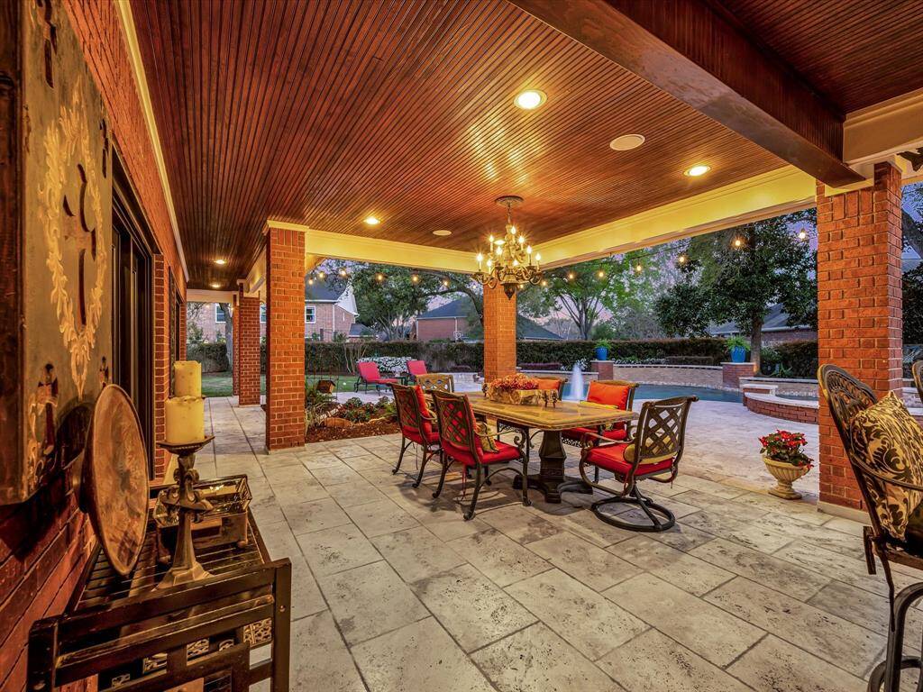 This photo showcases a beautiful covered patio with a wooden ceiling and brick pillars, featuring a stylish dining area with a chandelier. It overlooks a well-maintained backyard with a pool, perfect for outdoor entertaining.