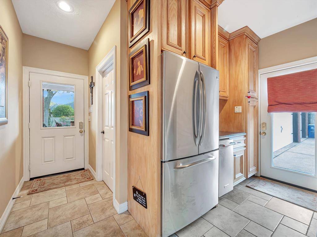 This photo showcases a cozy entryway leading into a kitchen area or to the backyard.  The space features light beige walls, tiled flooring, and wooden cabinetry. A stainless steel refrigerator and stand-alone icemaker are prominently placed, and there are two doors—one leading outside with a red window shade, allowing natural light to filter in. The area is both functional and welcoming.