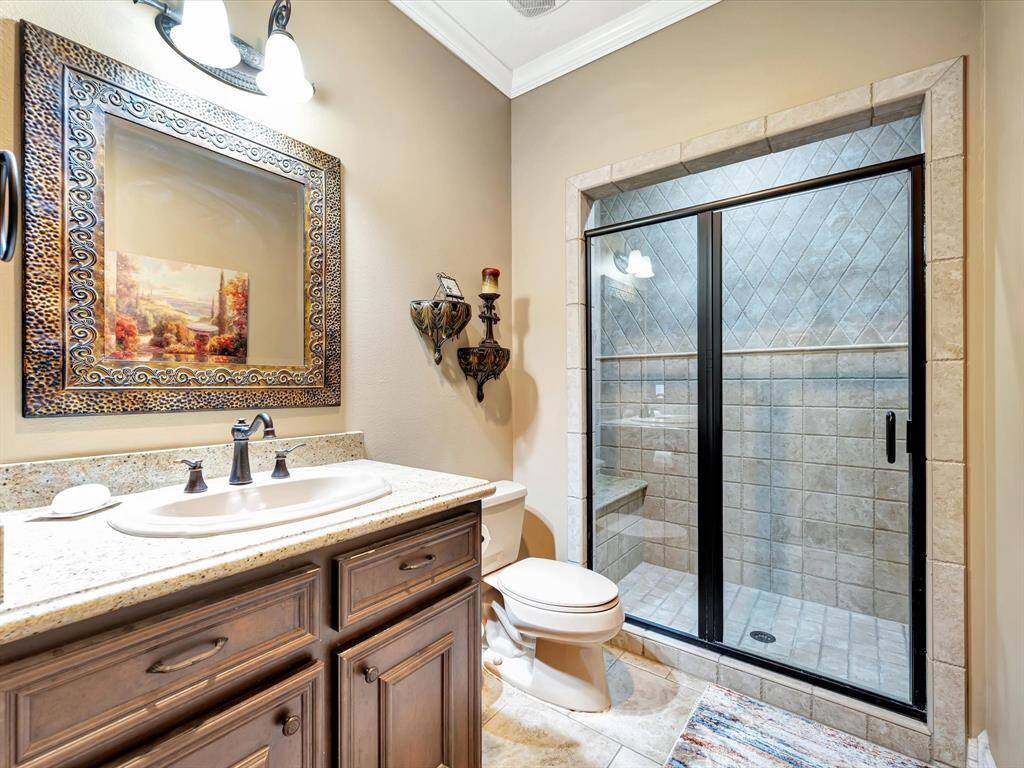 This downstairs bathroom features a spacious glass-enclosed shower with tile detailing, a granite countertop with a sink, and elegant wooden cabinetry. A large decorative mirror and tasteful lighting add a touch of luxury. It is located near the outdoor area.