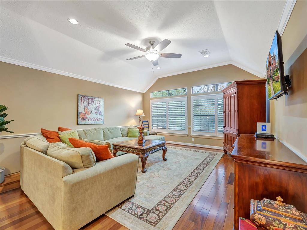 This bright media room features a vaulted ceiling with a fan, hardwood floors, and large windows with plantation shutters. It includes a cozy sectional sofa, a central coffee table, and a wall-mounted TV, making it ideal for relaxation and entertaining.
