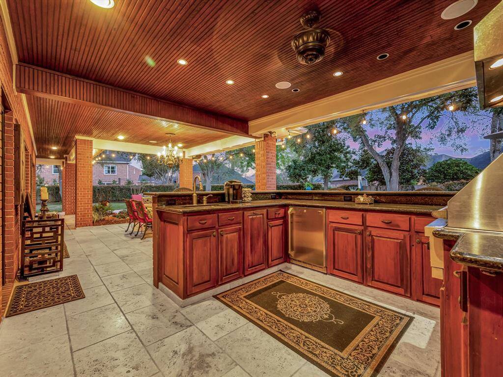 This photo showcases a luxurious outdoor kitchen and dining area with elegant wooden cabinetry, granite countertops, and ample lighting. The covered space features a ceiling fan, intricate tile flooring, and a scenic view of trees, perfect for entertaining.