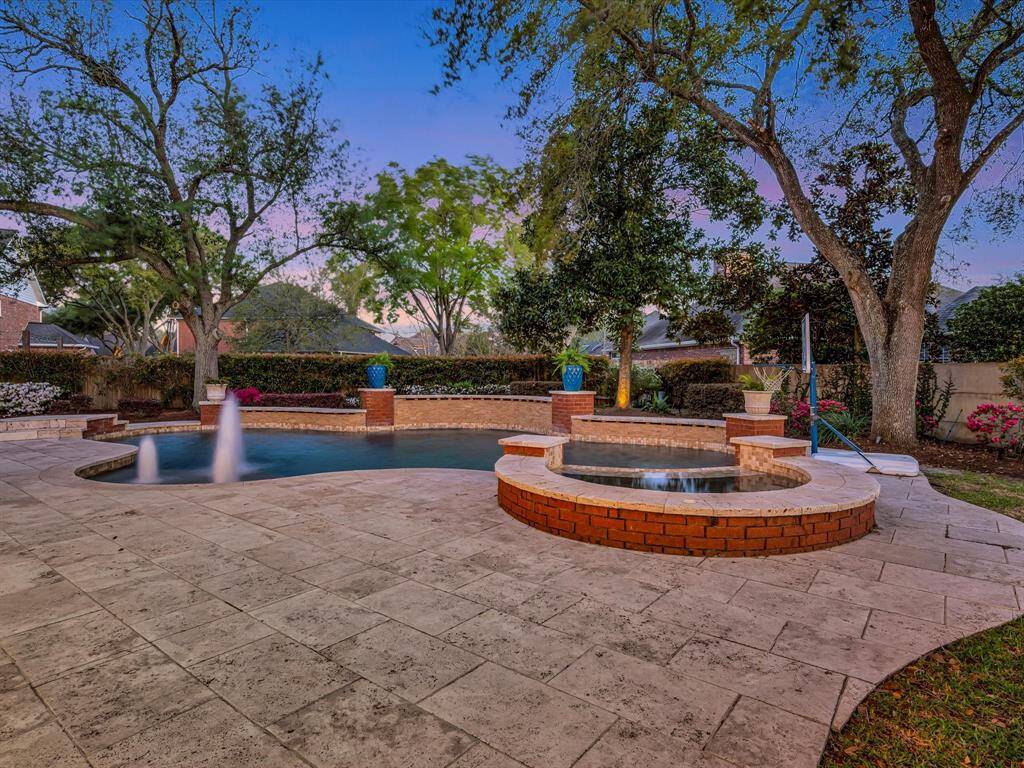 This photo showcases a serene backyard with a beautifully designed pool and spa area, surrounded by lush landscaping and mature trees. The stone patio provides ample space for relaxation and outdoor activities, creating an inviting oasis atmosphere.
