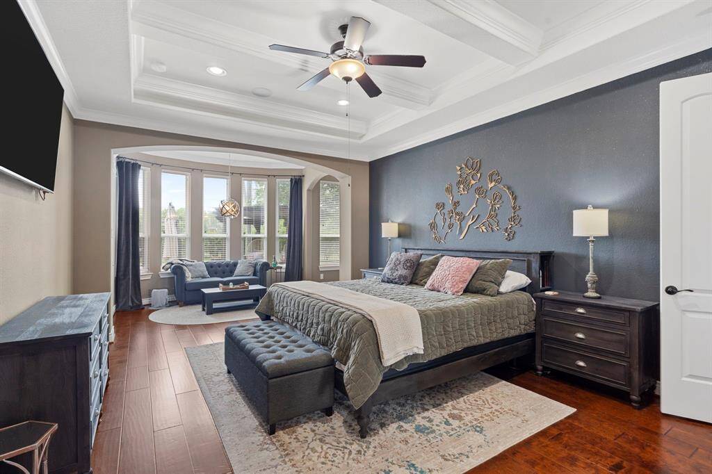 Luxurious primary suite featuring rich wood floors, a coffered ceiling, and a bright sitting area framed by a wall of windows—ideal for relaxing in style.