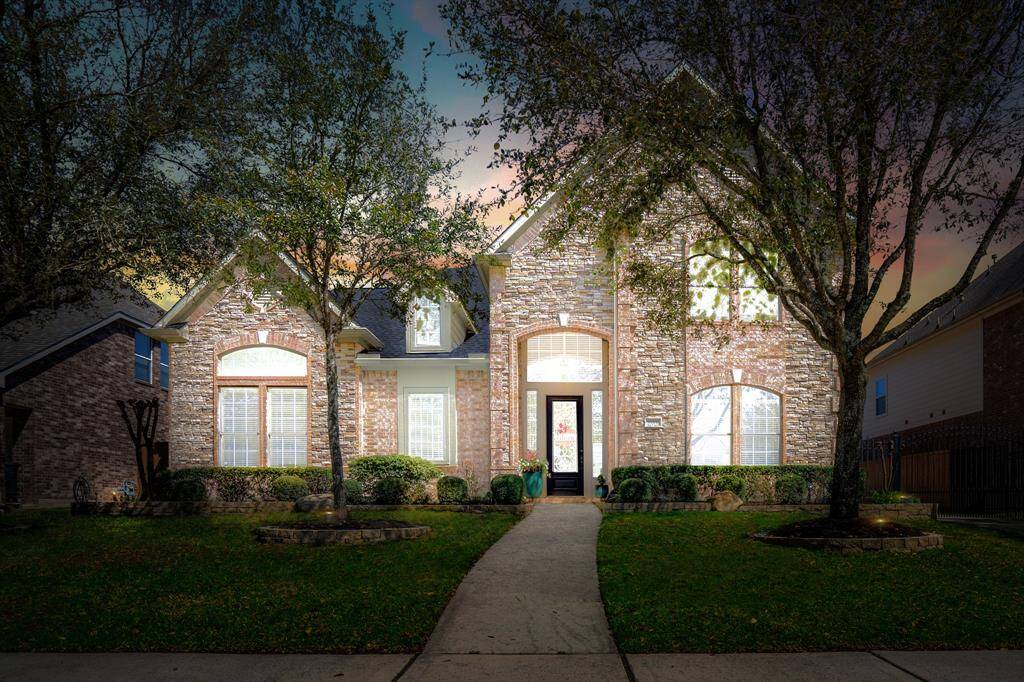 This stunning brick and stone-front home exudes curb appeal with mature trees, meticulous landscaping, and a grand arched entryway. A charming front walkway leads to the inviting entrance, while the warm interior glow contrasts beautifully against the vibrant sunset sky. Nestled in the sought-after Gleannloch Farms community, this home offers a perfect blend of luxury, sophistication, and suburban charm.