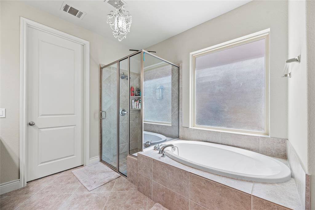 This bathroom features a spacious, elegant design with a glass-enclosed shower and a large soaking tub. Natural light streams through the frosted window, highlighting the tiled floor and walls. A modern chandelier adds a touch of luxury.