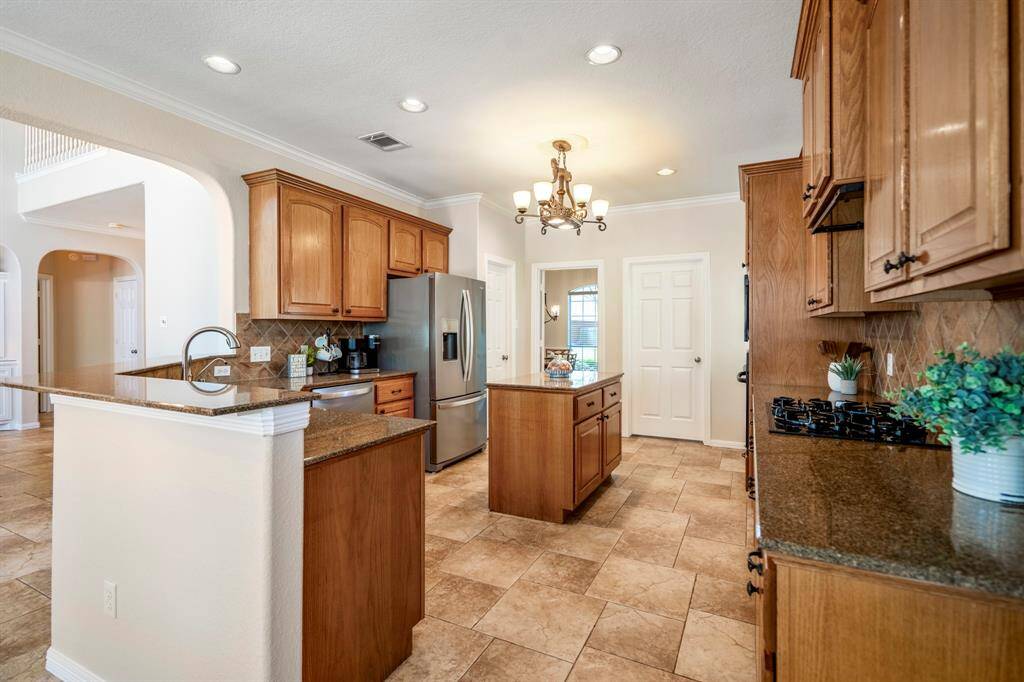 Moving closer into the kitchen, the spacious layout becomes even more evident. An elegant chandelier hangs over the center island, which offers plenty of storage. To the left, the kitchen features abundant counter space with a raised eating bar, a sink with a pull-out faucet, and a doorway leading to a massive walk-in pantry just to the right of the fridge. On the right side, you'll find the gas cooktop, built-in microwave, and oven. The doorway to the right leads to the laundry room.