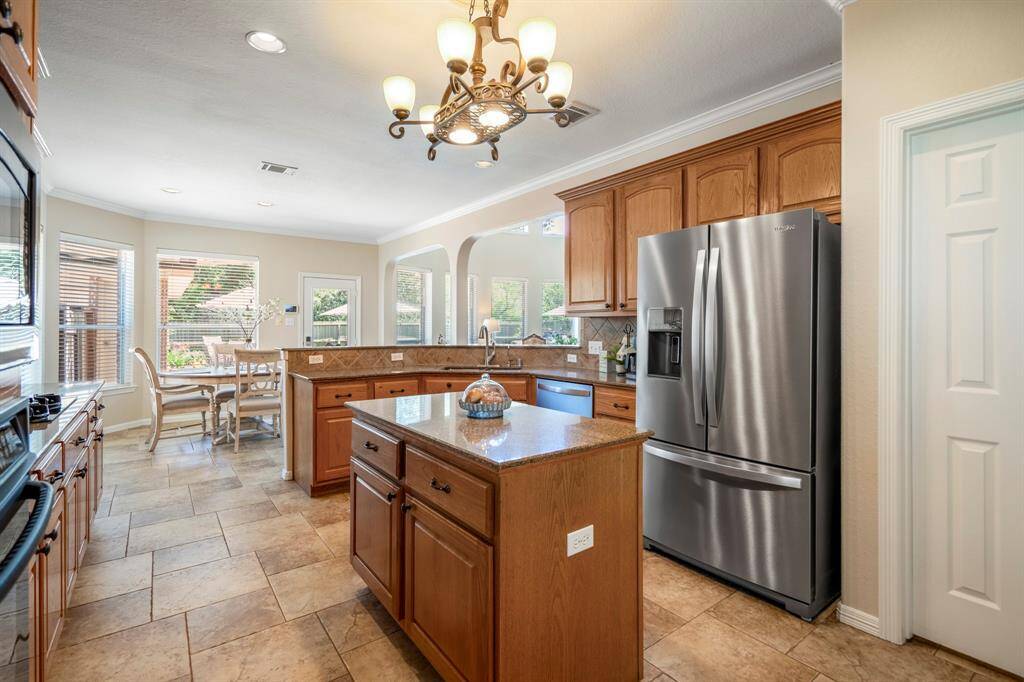 Taken from near the oven, this view looks back across the kitchen toward the breakfast area, offering a better glimpse of the space near the sink and fridge. The doorway to the massive walk-in pantry is visible to the right, providing easy access to additional storage. Beautiful wood cabinets with detailed trim work, granite counters, and a tile backsplash add both style and function to this well-appointed kitchen.