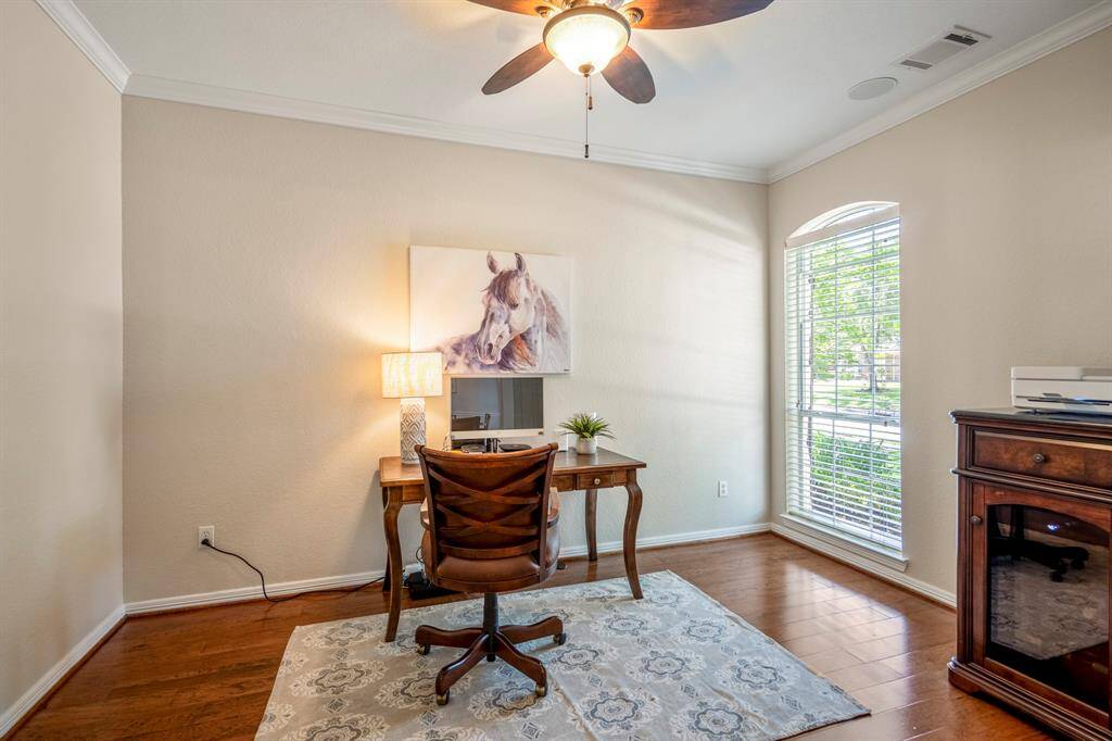 A peek inside the home office reveals a spacious workspace with gleaming wood floors, a large window offering views of the front yard, a ceiling fan for added comfort, and elegant crown molding, creating a perfect environment for productivity.