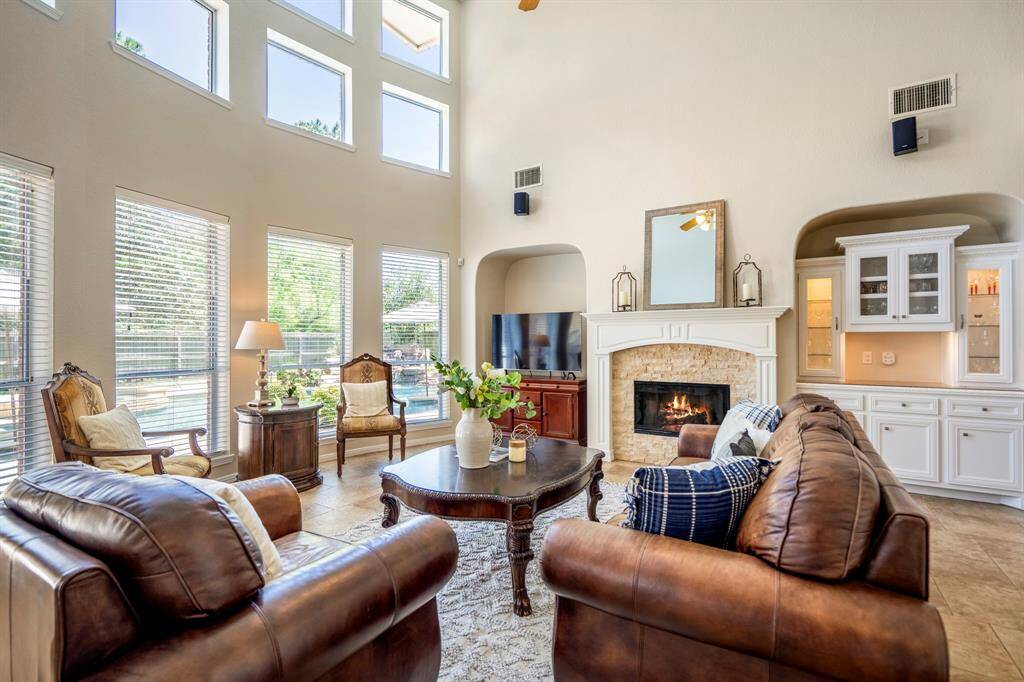 Stepping into the living room, you'll be captivated by the stunning wall of windows along the back of the home, offering picturesque views of the pool and flooding the space with natural light. The two-story ceilings and windows extend all the way up, creating a dramatic, airy atmosphere. The gas log fireplace, with its stacked stone surround and elegant wood-trimmed mantle, serves as a beautiful focal point. Recessed archways to either side provide a spot for the TV on the left and built-in cabinets.