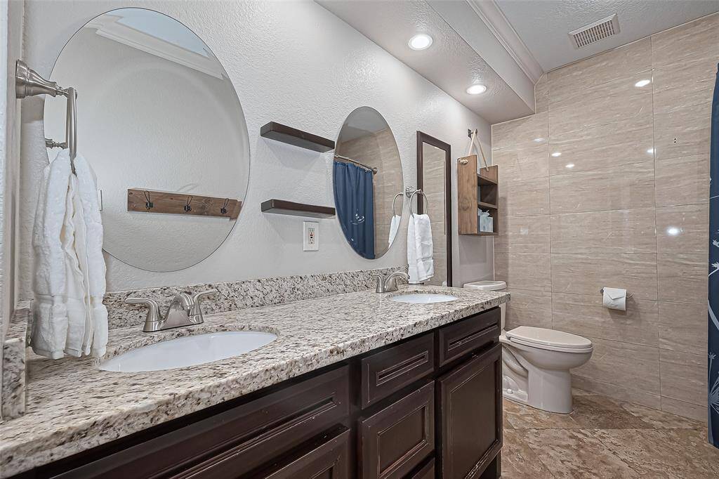 Step into this beautifully designed bathroom featuring a granite-topped double sink vanity—perfect for busy mornings or shared routines—with modern round mirrors, soft lighting, and elegant tile work that extends into the shower for a clean, polished finish.